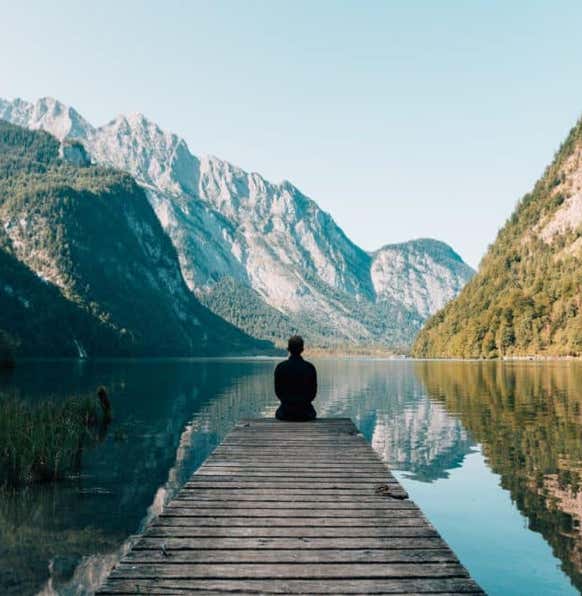 une personne de dos assise au bout d'un ponton faisant face à un lac dans lequel se reflète le paysage de montagne en arrière plan, illustrant le dalme et la sérénité grâce au massage métamorphique - cabinet Terre de Shiatsu Lyon 5e
